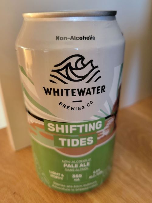 A can of Whitewater Brewing Co. Shifting Tides Non-Alcoholic Pale Ale. The can is silver with a green and blue design featuring a wave and a mountain. The text "Shifting Tides" is prominently displayed in a green font. The can is 355ml and contains 0% alcohol.
