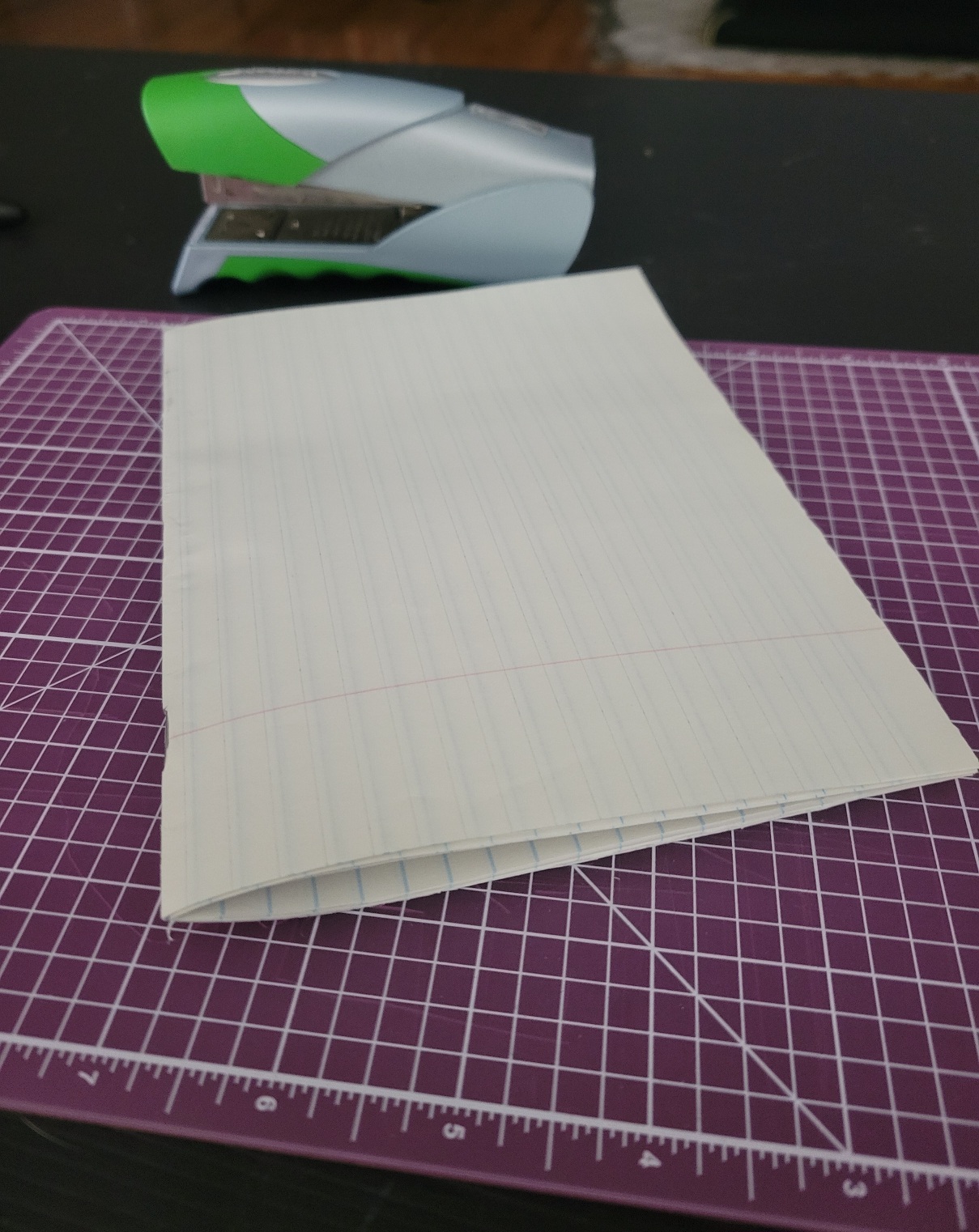 A lined sheet of paper folded in half, resting on a purple cutting mat with grid lines. A green and silver stapler is in the background on a dark surface.
