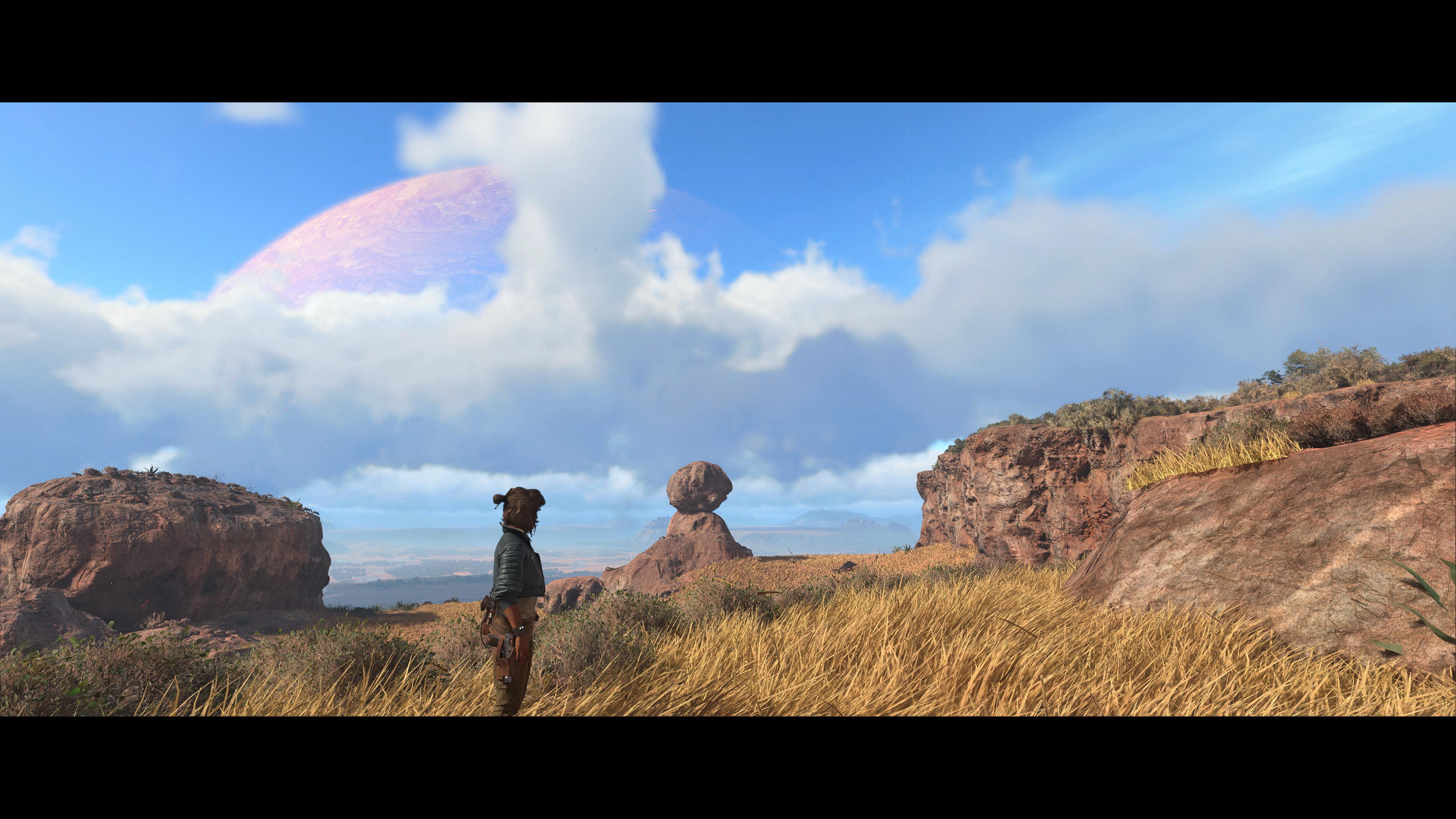 A panoramic screenshot from the game Star Wars Outlaws showing the protagonist, Kay Vess, standing in tall, dry grass on a rocky alien planet. In the foreground, there are arid shrubs and golden grass. Rocky cliffs flank the scene, and a naturally balanced, mushroom-shaped rock formation stands in the middle distance. A massive, reddish-pink ringed planet dominates the sky through scattered white clouds. Alt text generated by Google Gemini.