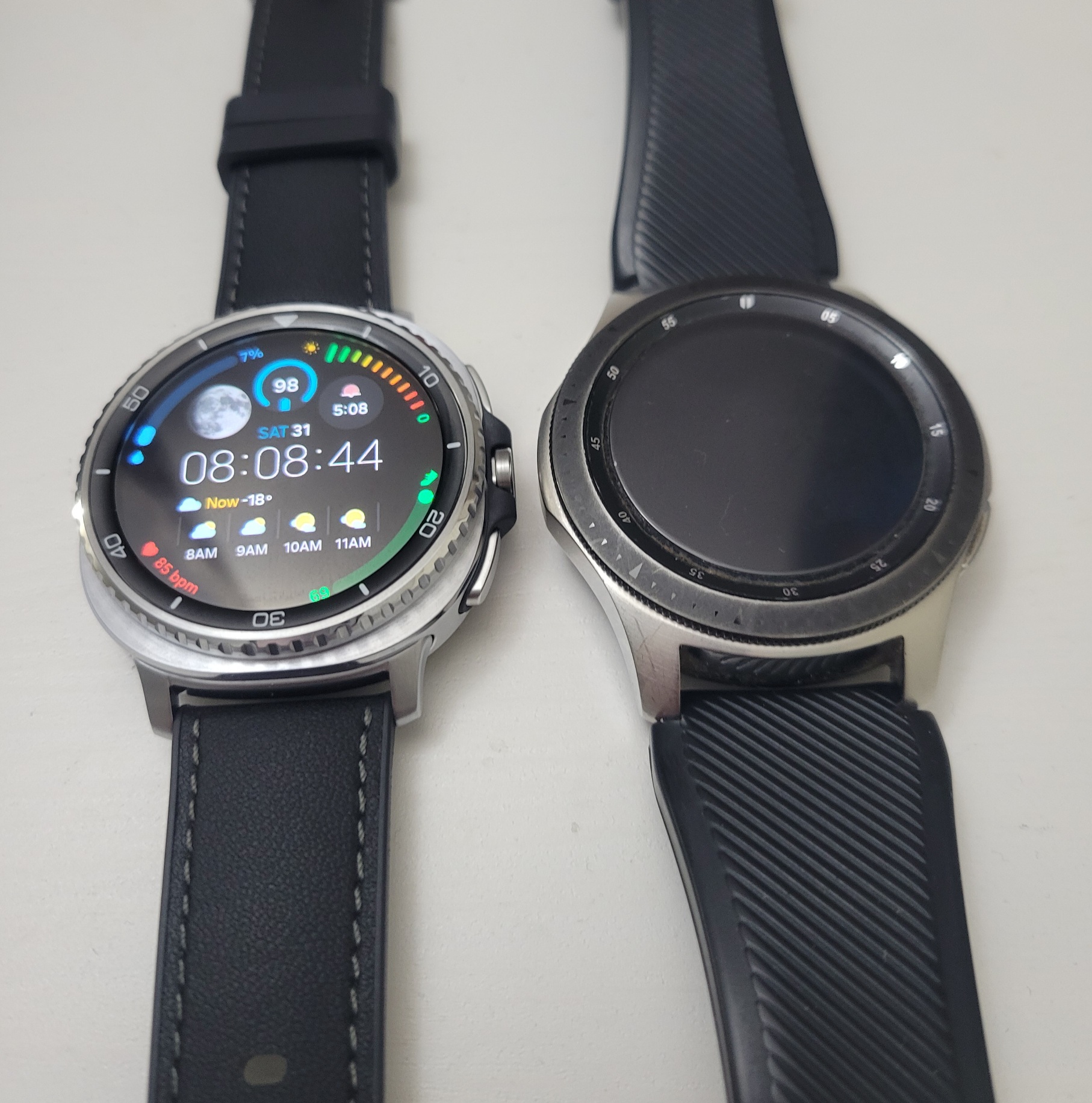 Two smartwatches are lying on a surface beside each other. On the left, a Samsung Galaxy Watch 8 Classic, and on the right a Samsung Galaxy Watch 46MM. The watch on the left is displaying a digital watch face with various statistics. The time reads 8:08:44.